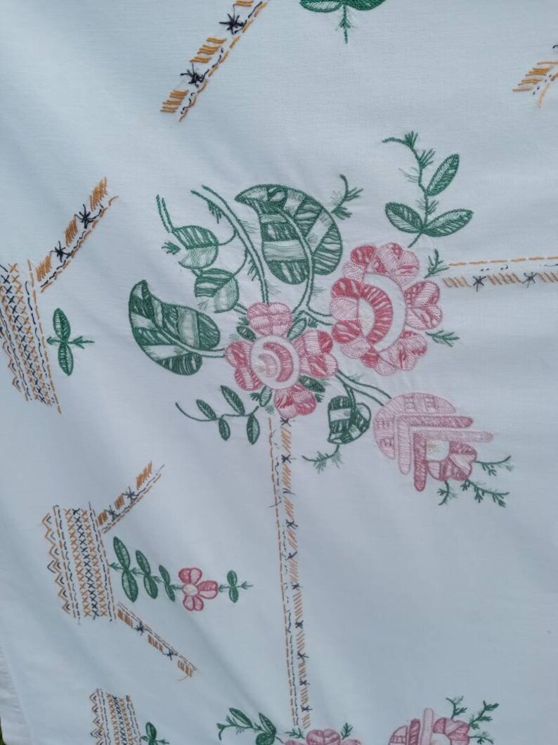 May include: White tablecloth with an embroidered floral design. The flowers are pink and green with a woven pattern. The tablecloth has a border of brown and gold stitching.