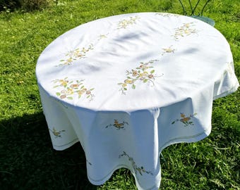 Könnte beinhalten: Runder weißer Tischdecken mit gestickten Blumendesigns in Gelb-, Grün- und Brauntönen. Die Tischdecke liegt auf einem Tisch in einer grasbewachsenen Außenumgebung.