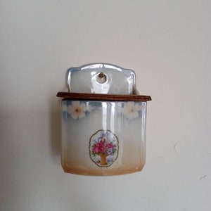 May include: A white ceramic wall-mounted matchbox holder with a floral design. The holder has a wooden top and a small hole in the front.
