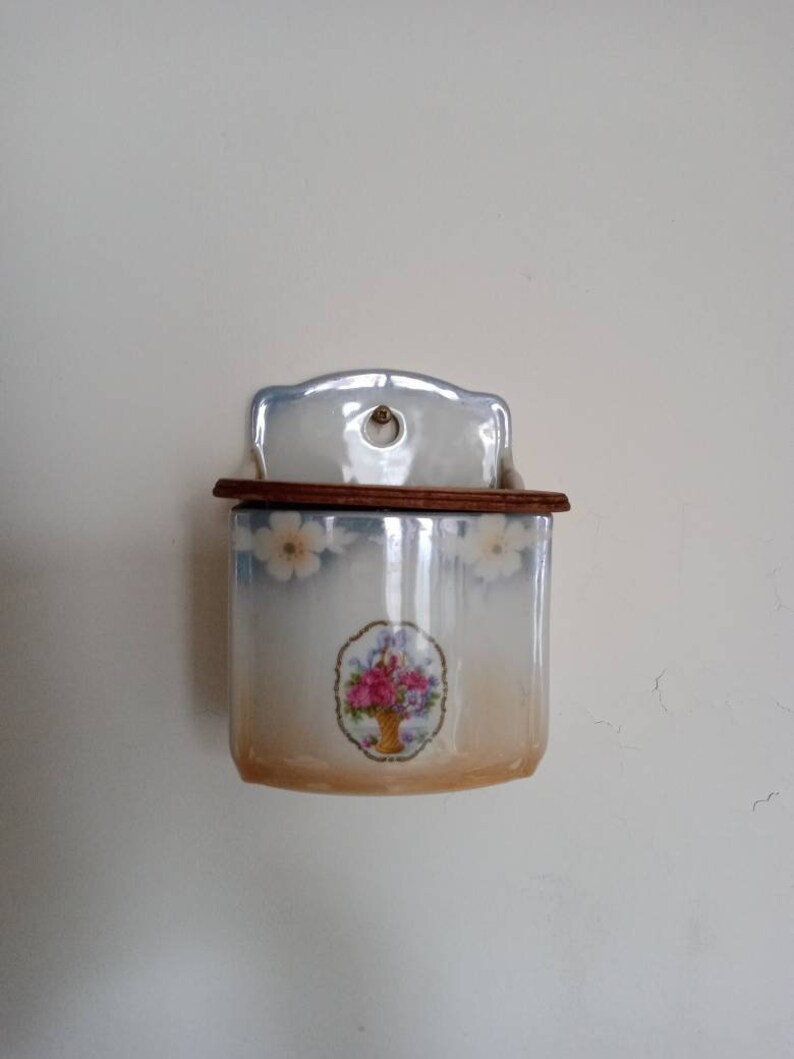 May include: A white ceramic wall-mounted matchbox holder with a floral design. The holder has a wooden top and a small hole in the front.