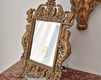 May include: A gold-toned ornate mirror with a decorative frame featuring cherub figures and intricate scrollwork. The mirror is standing on a marble surface.
