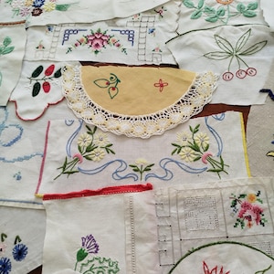 Op de afbeelding: Een verzameling diverse witte en gebroken witte stoffen met verschillende kleurrijke bloemenborduursels, kruissteekpatronen en delicate kanten randen. De textielstukken tonen diverse botanische motieven, waaronder kersen, bloemen en linten in tinten rood, geel, blauw, groen en paars.