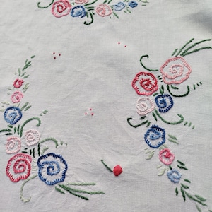 May include: White linen tablecloth featuring embroidered floral designs in pink, blue, and green. The flowers are arranged in a circular pattern.