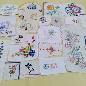 Op de afbeelding: Een verzameling diverse witte stoffen vierkanten en rechthoeken met verschillende kleurrijke bloemen-, vogel- en botanische borduurontwerpen, waaronder enkele stukken met kanten randen, schulpranden en de geborduurde letters CUC.