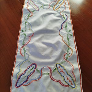 Peut inclure: Un chemin de table blanc avec une bordure brodée colorée. La bordure présente un motif répétitif de feuilles stylisées dans des tons de rouge, bleu, vert et jaune.