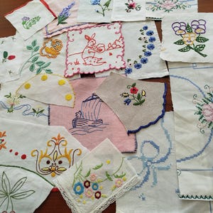 Puede incluir: Una colección de cuadrados de tela blancos y de colores variados con diversos diseños bordados a mano, que incluyen flores, un conejo, un velero y patrones geométricos. Las piezas varían en forma, algunas con bordes festoneados o de encaje.