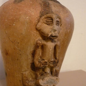 Può includere: Un vaso in terracotta con un viso e un corpo umani scolpiti. Il vaso è marrone e ha una texture ruvida.