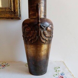 Op de afbeelding: Een hoge, bronzen vaas met een getextureerd oppervlak en een ontwerp van bladeren en bessen. De vaas heeft twee handvatten en een licht uitlopende opening.