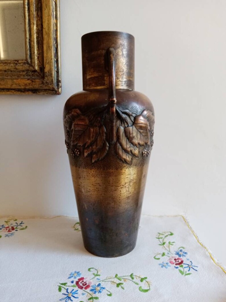 Op de afbeelding: Een hoge, bronzen vaas met een getextureerd oppervlak en een ontwerp van bladeren en bessen. De vaas heeft twee handvatten en een licht uitlopende opening.