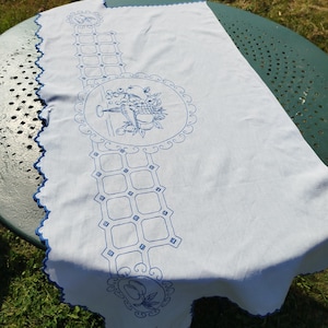 May include: White linen table runner with blue embroidered designs. The design features a repeating pattern of squares and circles with fruit and floral motifs.