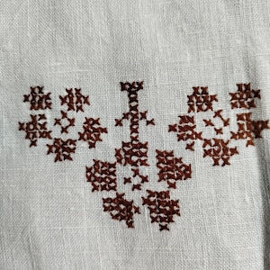 May include: A white linen tea towel with a brown embroidered design of a stylised flower with a central stem.