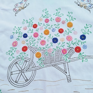 Op de afbeelding: Wit geborduurd tafelloper met een zwarte kruiwagen gevuld met kleurrijke bloemen in rood, geel, roze en blauw. Twee vlinders zijn bovenaan geborduurd. De loper heeft een groene rand.