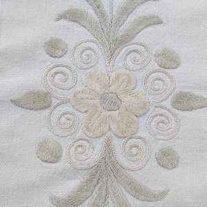 Pode incluir: Tecido branco com um desenho floral bordado. O desenho apresenta duas flores com um caule e folhas. As flores são bege e as folhas são verde claro.