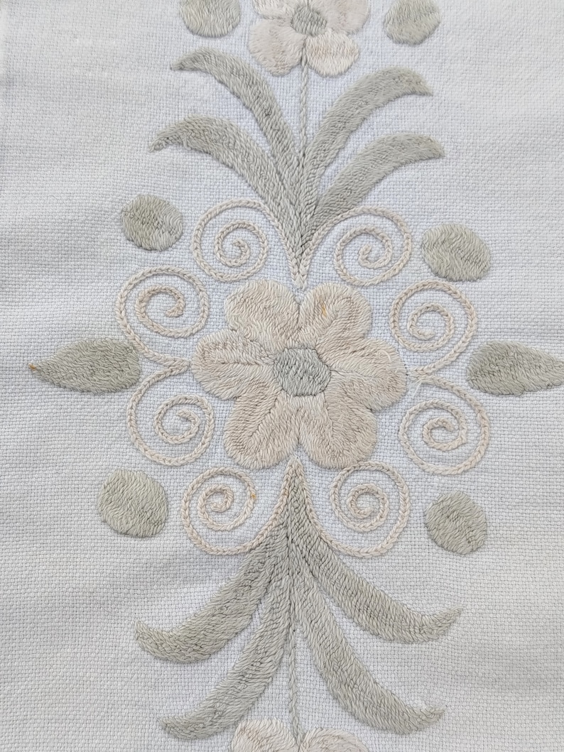 May include: White fabric with embroidered floral design. The design features two flowers with a stem and leaves. The flowers are beige and the leaves are a light green.