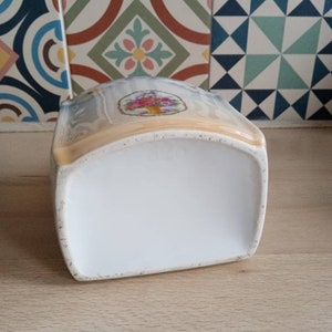 May include: A white ceramic jar with a floral design on the top. The jar has a rounded shape and a smooth, white finish.