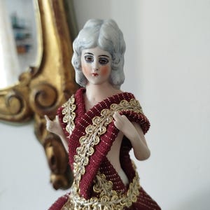Op de afbeelding: Een porseleinen pop met een bleke huid, geschilderde gelaatstrekken en golvend wit haar. De pop draagt een kledingstuk van gestructureerde donkerrode stof dat over de schouders is gedrapeerd, geaccentueerd met gouden metallic randen en gouden kant.