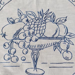 May include: A blue embroidered design of a fruit bowl filled with grapes, apples, and a pineapple. The bowl is on a table with a white linen background.