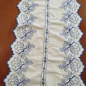 May include: A white table runner featuring blue embroidered cherry motifs along both scalloped edges. A central vertical band displays a repeating blue floral pattern. The fabric has a clean, light-colored appearance with detailed blue stitching throughout.