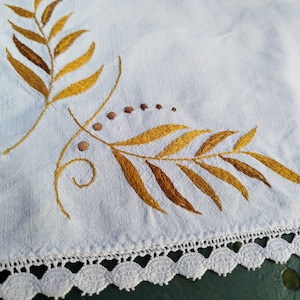 May include: White linen tablecloth with a gold embroidered leaf design. The tablecloth has a white crocheted border.