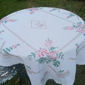 May include: White tablecloth with embroidered floral designs in pink, green, and brown. The tablecloth is laid on a round table with a green metal chair in the background.