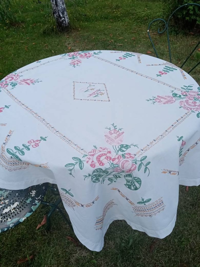 May include: White tablecloth with embroidered floral designs in pink, green, and brown. The tablecloth is laid on a round table with a green metal chair in the background.
