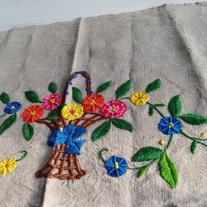 May include: Embroidered linen fabric with a brown wicker basket filled with colorful flowers. The flowers are red, yellow, blue, and pink. The basket is surrounded by green leaves and vines.