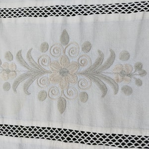 May include: White linen table runner with a delicate embroidered floral design in shades of gray. The runner features a central flower with swirling tendrils and smaller flowers on either side. The edges of the runner are trimmed with a white crochet lace.