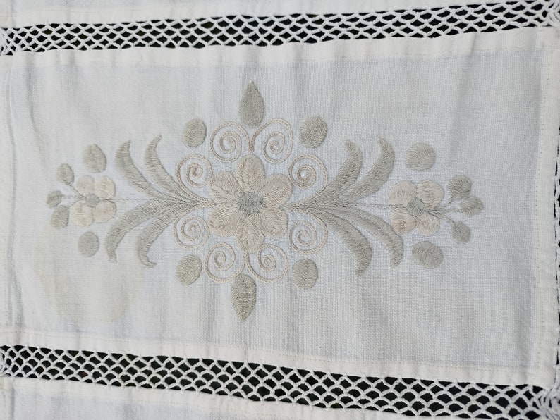 May include: White linen table runner with a delicate embroidered floral design in shades of gray. The runner features a central flower with swirling tendrils and smaller flowers on either side. The edges of the runner are trimmed with a white crochet lace.