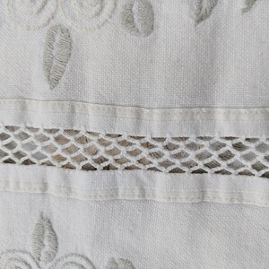 May include: White fabric with a delicate embroidered design featuring floral motifs and a lace trim. The fabric has a subtle texture and a soft, white color.