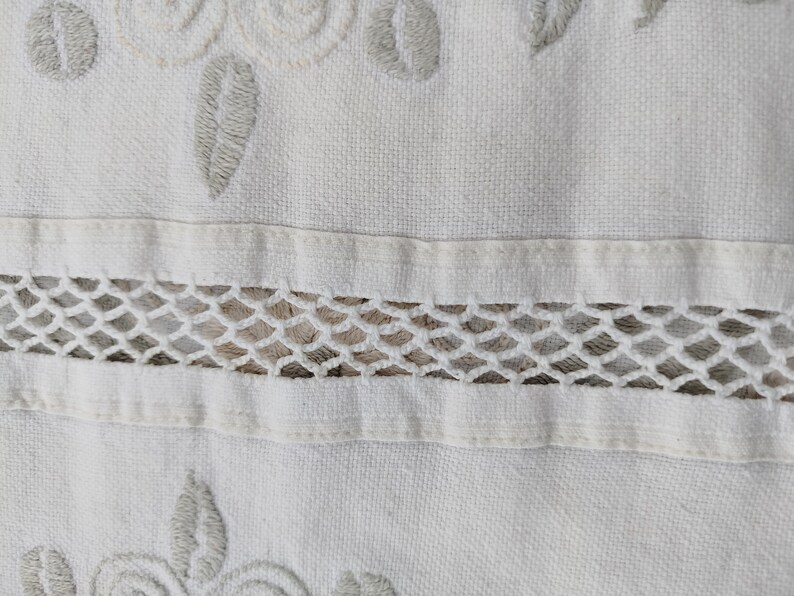 May include: White fabric with a delicate embroidered design featuring floral motifs and a lace trim. The fabric has a subtle texture and a soft, white color.
