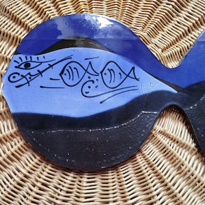 Könnte beinhalten: Keramik-Fischplatte in Blau und Schwarz. Die Platte hat ein stilisiertes Design von Fischen und anderen abstrakten Elementen in Schwarz. Die Platte liegt auf einer gewebten Korbweidenoberfläche. Die Signatur des Künstlers ist sichtbar.