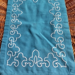 May include: A turquoise blue fabric placemat with a white embroidered border. The border features a repeating pattern of stylized floral designs.