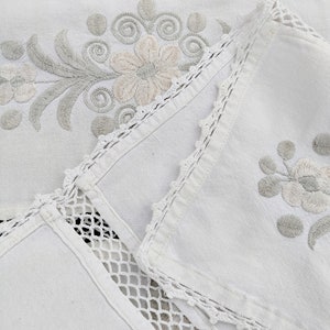May include: White linen tablecloth with delicate floral embroidery and crocheted trim. The tablecloth is folded in half, revealing a second layer of fabric with a similar design.