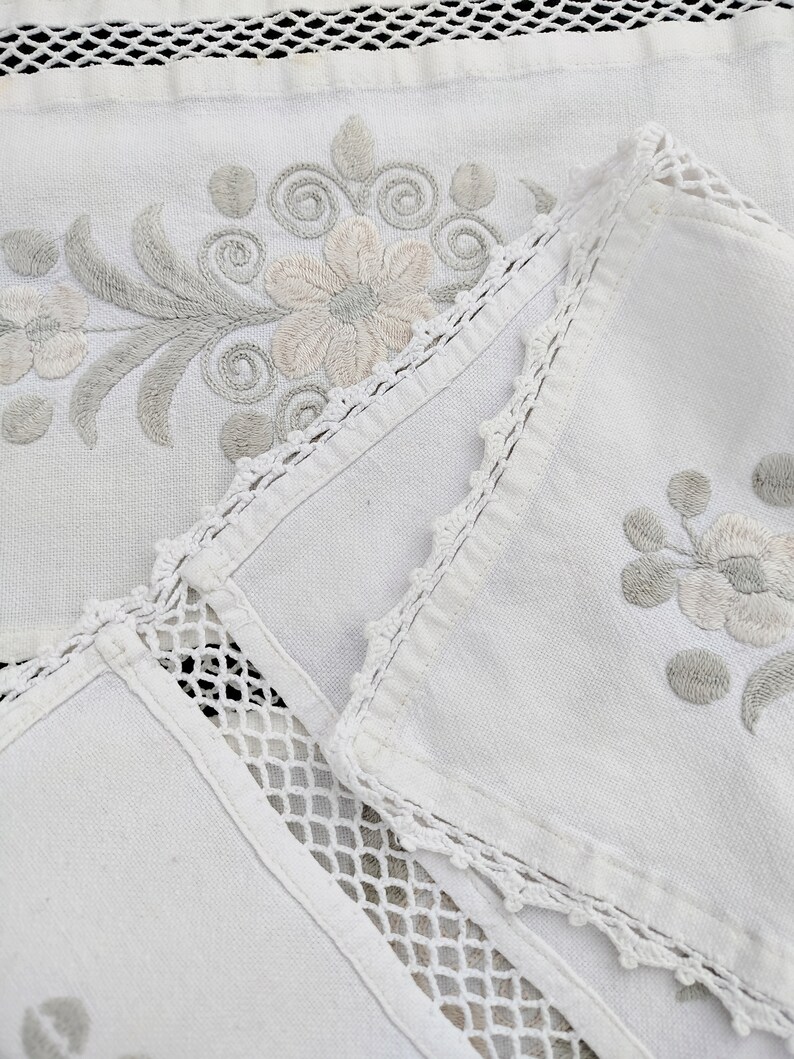 May include: White linen tablecloth with delicate floral embroidery and crocheted trim. The tablecloth is folded in half, revealing a second layer of fabric with a similar design.