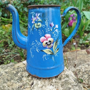 Può includere: Una teiera blu in smalto con beccuccio e manico curvi. La teiera è decorata con disegni floreali dipinti a mano in rosa, viola e bianco. Il bordo della teiera ha un colore marrone ruggine.