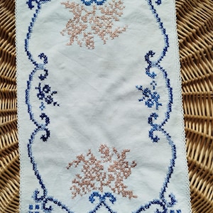 May include: White embroidered cloth with a blue border and floral designs. The border has a delicate lace edge. Two clusters of peach-coloured floral embroidery are centered on the cloth.