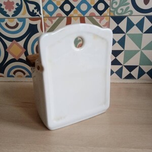 May include: White ceramic toothbrush holder with a rounded top and a hole in the back for hanging. The holder has a wooden base.