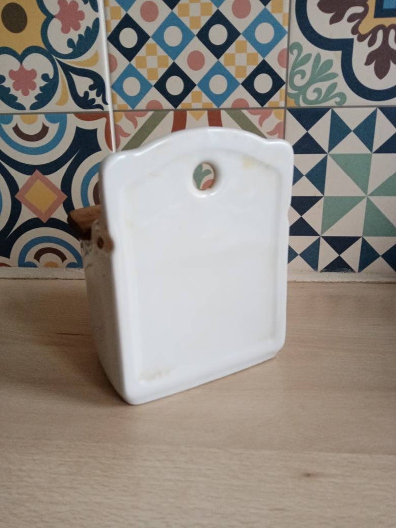 May include: White ceramic toothbrush holder with a rounded top and a hole in the back for hanging. The holder has a wooden base.