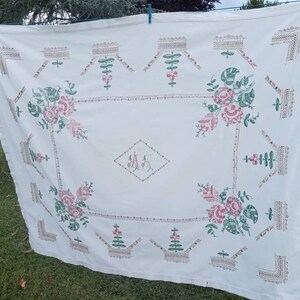 May include: White linen tablecloth with embroidered floral designs in shades of pink and green. The tablecloth features a central monogram "A" in a diamond shape.