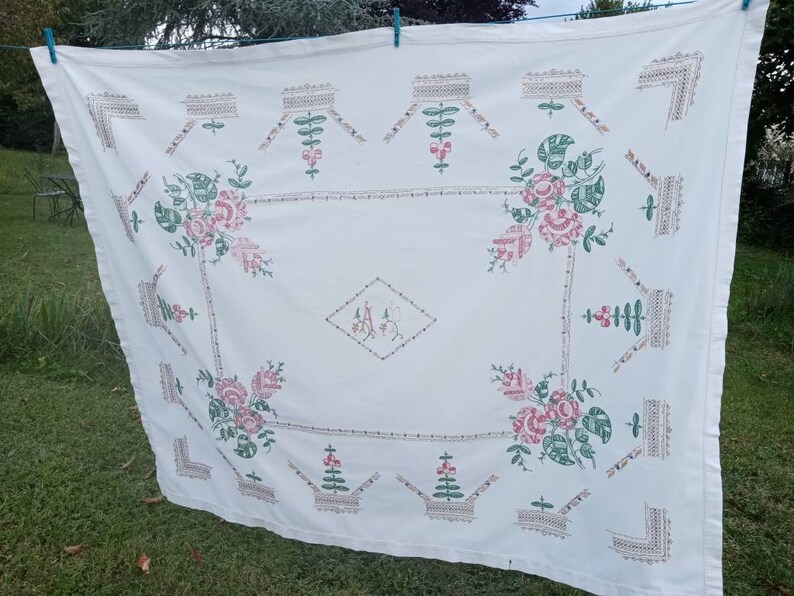 May include: White linen tablecloth with embroidered floral designs in shades of pink and green. The tablecloth features a central monogram "A" in a diamond shape.