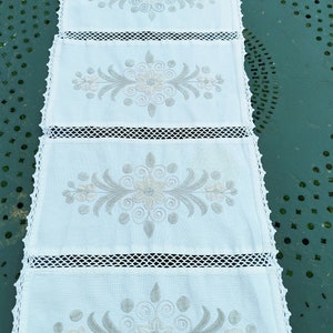 May include: White crocheted table runner with five embroidered floral designs in gray. The runner has a delicate lace trim.