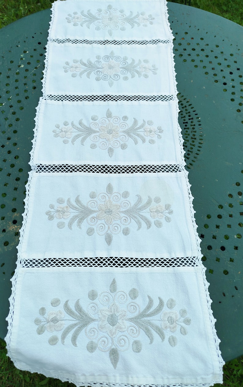 May include: White crocheted table runner with five embroidered floral designs in gray. The runner has a delicate lace trim.