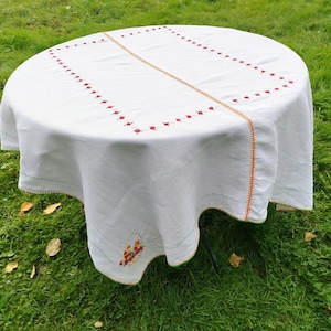 Peut inclure: Nappe en lin blanc avec des détails brodés rouges et orange. La nappe a un bord festonné et présente un petit panier de fleurs brodé au centre.