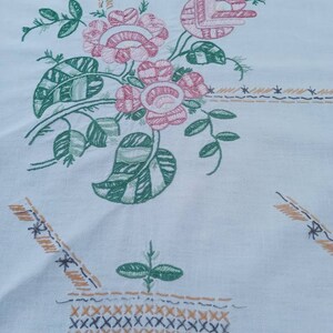 May include: White tablecloth with an embroidered floral design. The design features pink and green flowers with leaves, and a basket with a brown and black stitch pattern.