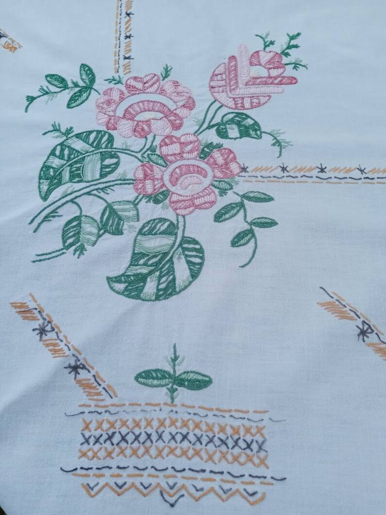 May include: White tablecloth with an embroidered floral design. The design features pink and green flowers with leaves, and a basket with a brown and black stitch pattern.