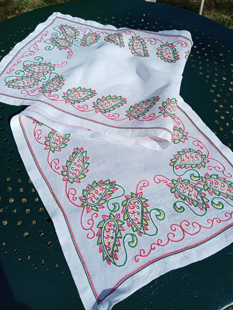 May include: White table runner with a paisley pattern embroidered in red, green, yellow, and purple thread.