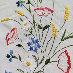 May include: White linen tablecloth with embroidered flowers in shades of red, white, blue, and yellow. The flowers include poppies, daisies, and cornflowers.