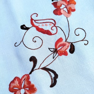 May include: Embroidered floral design on a light blue fabric. The design features three red and pink flowers with brown stems and leaves.