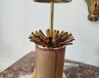 May include: A gold-toned metal cigarette dispenser featuring a cylindrical body with a woven-textured tan surface. The top mechanism includes a central vertical rod topped with a circular lid, surrounded by a fan of individual metal slots.
