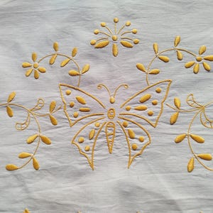 May include: Close-up of a light blue fabric with gold embroidery. The embroidery features a large butterfly in the center, surrounded by floral and leaf designs. The butterfly has detailed wing patterns.